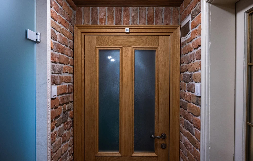 A wooden door with glass panes in a brick wall, with a small white device mounted on the upper door frame