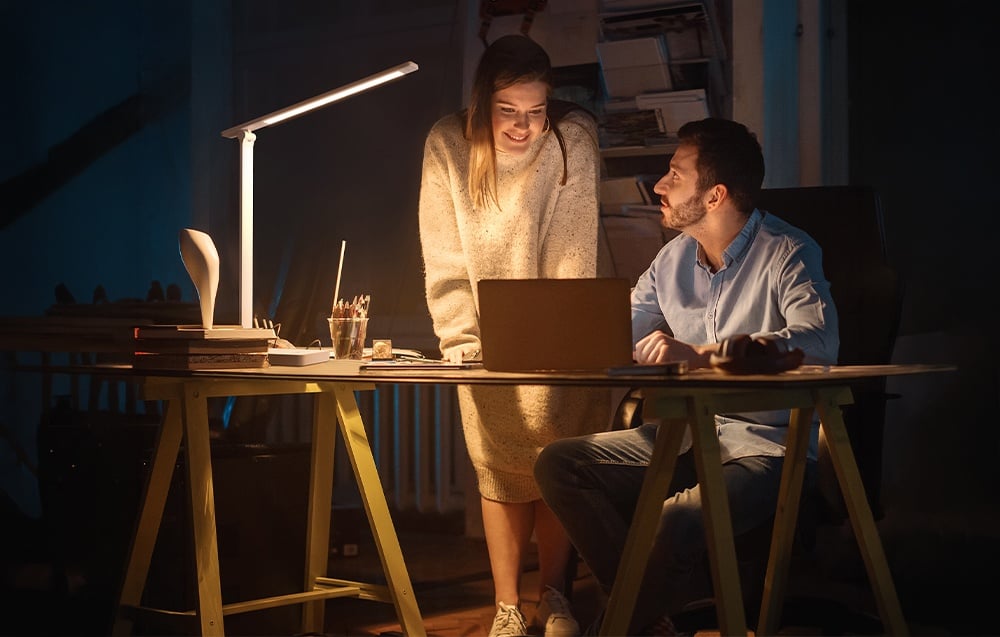 A woman and a man stand by a desk in a dark room, looking at a laptop screen illuminated by a white desk lamp.