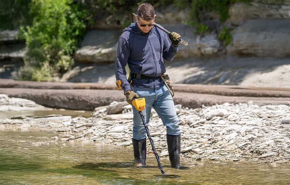 A man using a metal detector in shallow water, holding a paddle.