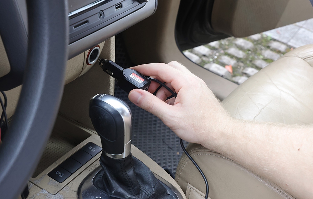 A man plugs a black power adapter into the cigarette lighter socket inside a brightly lit car