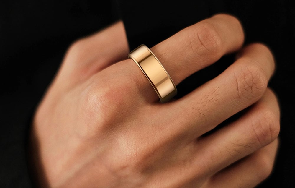 A man’s hand in a black jacket wearing a gold smart ring on his index finger