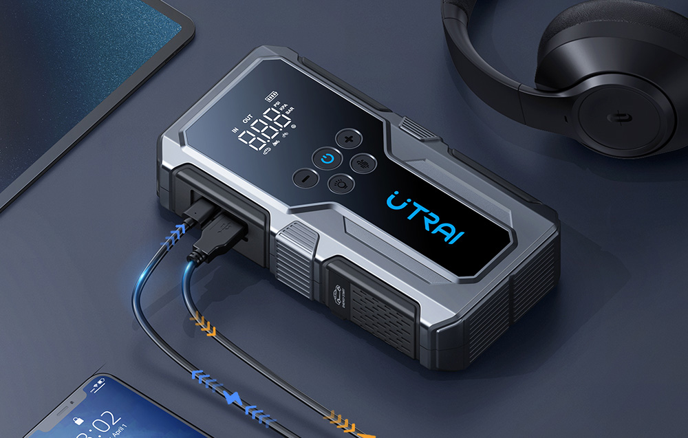A car jump starter lies on a table next to headphones, and the cables connected to it are charging a cell phone.