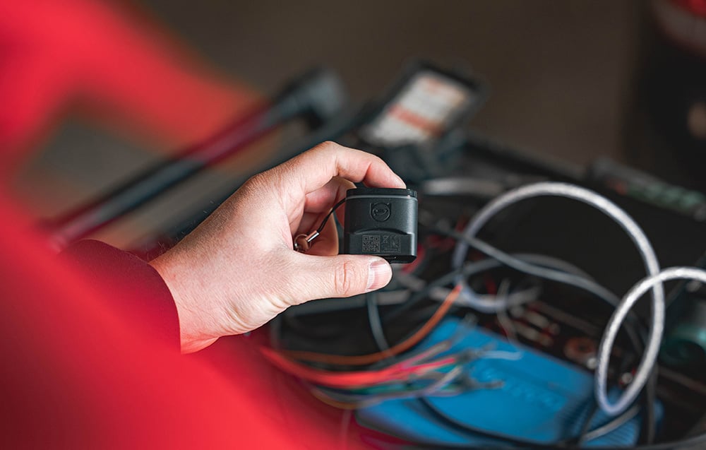 A hand in a red sweatshirt holding the Carly OBD2 adapter with the hood of an Audi car open