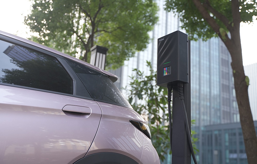 A light-colored car connected to an EV charger, with buildings in the background