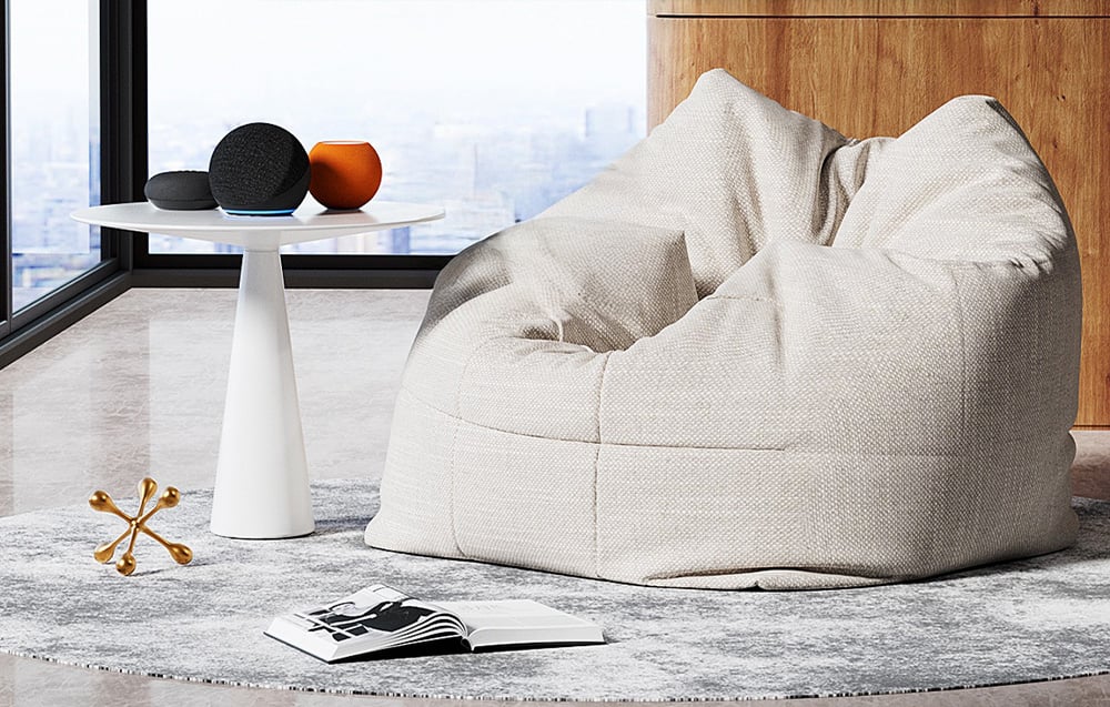 A gray pouf in the living room next to a white side table with decorations by a large window, an open book on the rug