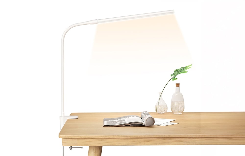 A white desk lamp clipped to the edge of a light-colored tabletop, next to an open newspaper and two vases with plants.
