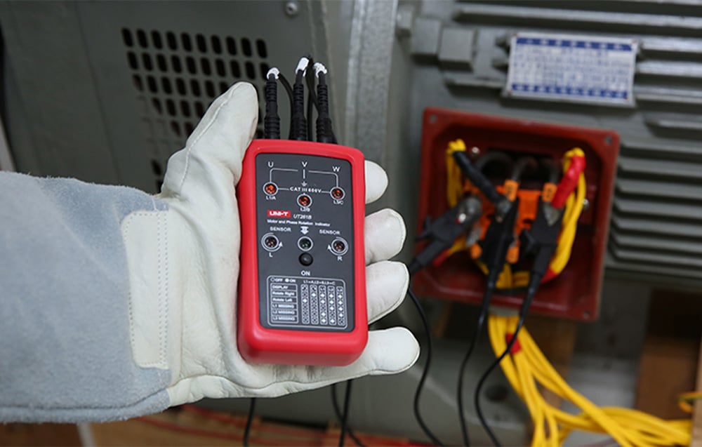A person wearing a glove holds the phase sequence tester connected to the terminals of the electric motor.