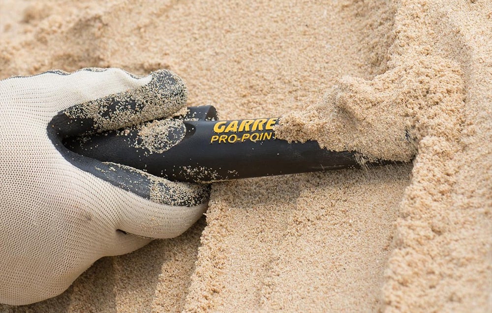 A gloved hand uses a metal detector in the sand.