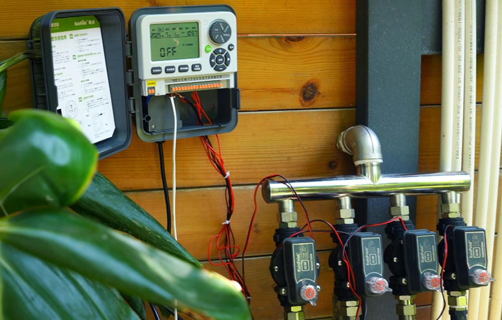 An open RainPoint® irrigation controller mounted on a wooden wall, connected by wires to four solenoid valves installed on a metal water manifold.
