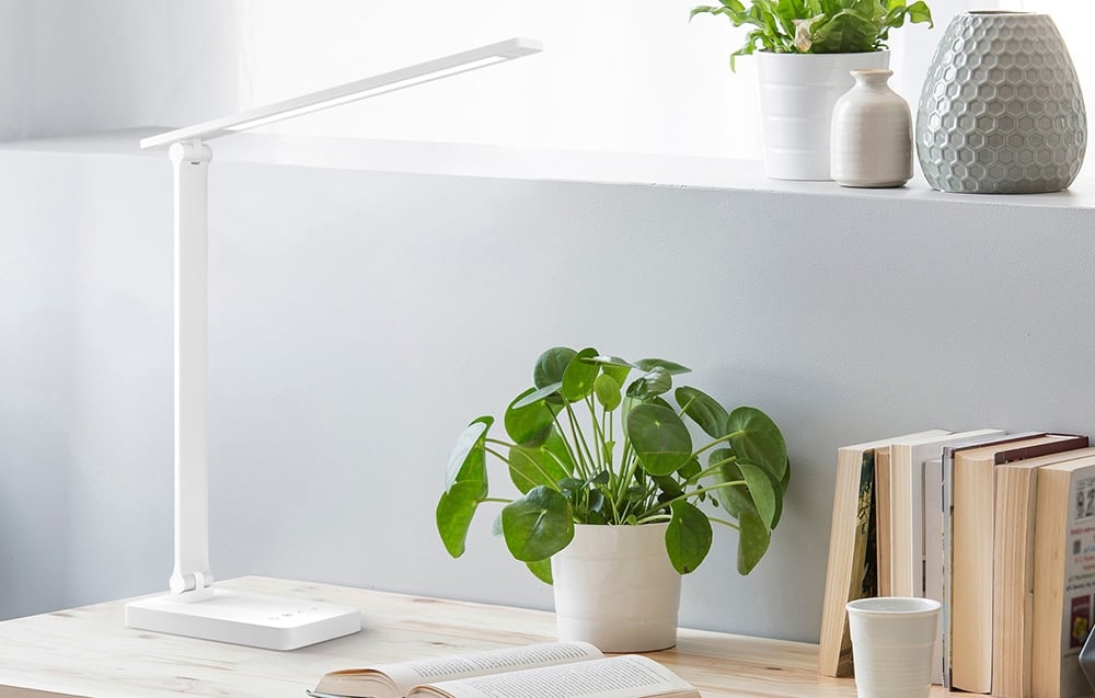 A white, elongated desk lamp stands on a light-colored wooden tabletop next to a green potted plant and books