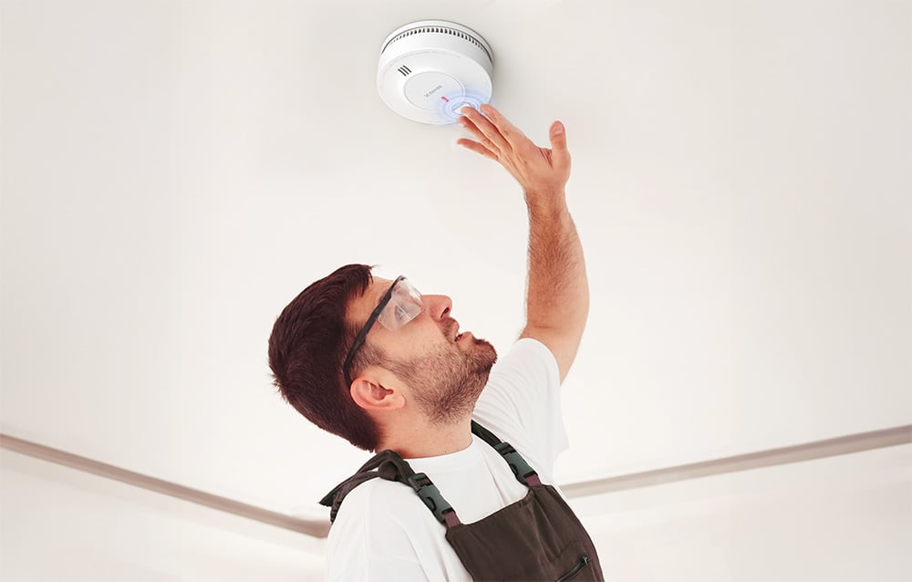 A man wearing safety goggles and a coverall inspects the smoke detector on the ceiling