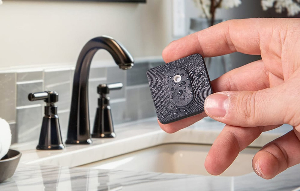 A hand holding a damp, black Shelly sensor against the backdrop of a sink and a modern bathroom faucet