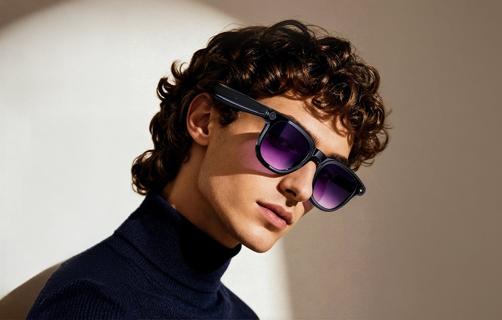 A man in a dark turtleneck and smart glasses with purple lenses looks to the side