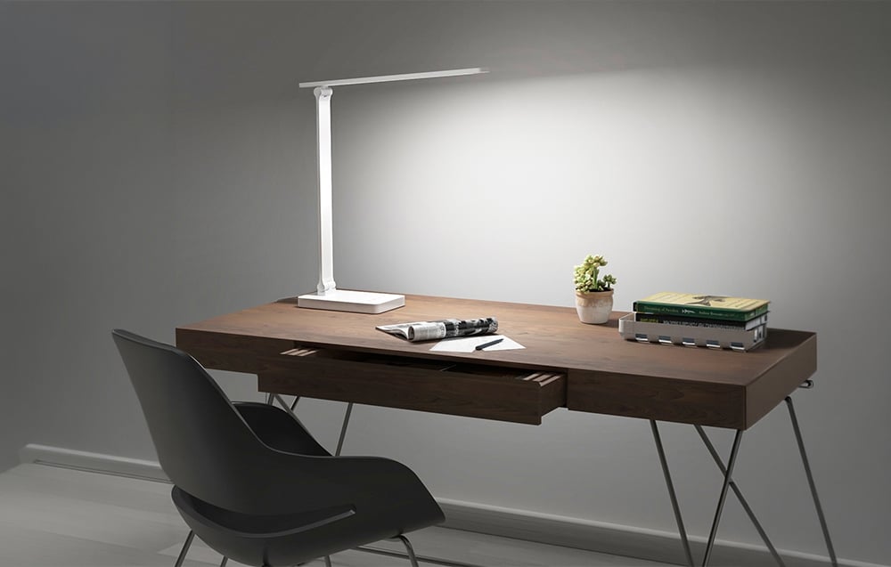 A modern, white desk lamp illuminates a wooden tabletop with an open newspaper, books, and a small plant.