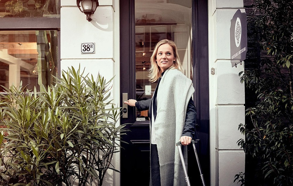 A woman with a suitcase and a light-colored scarf opens the dark front door of a building marked with the number 38H