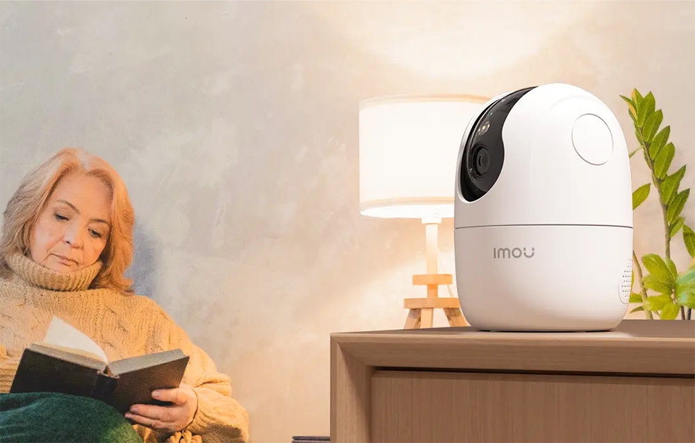 A white Imou camera on a cabinet, in the background a woman reading a book by a lit lamp