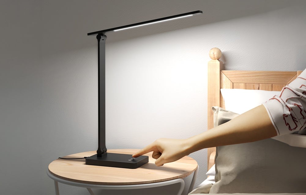A hand touches the touch panel on the base of a black, lit desk lamp standing by the bed.