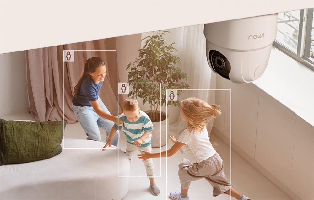 A white camera mounted on the ceiling monitors the living room, where two children and a woman are playing; the figures are framed in white.