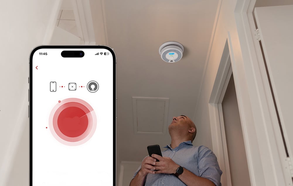 A man in the hallway looks at the smoke detector on the ceiling, holding a phone with the mobile app in the foreground