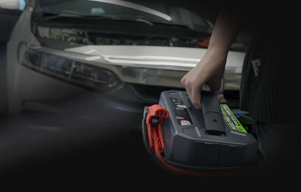 A person carrying the compact jump starter by its handle near the open hood of a white car.