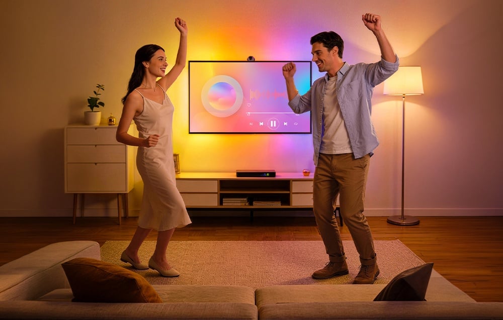 A smiling couple dances in the living room in front of the TV; the room is lit with colorful lights