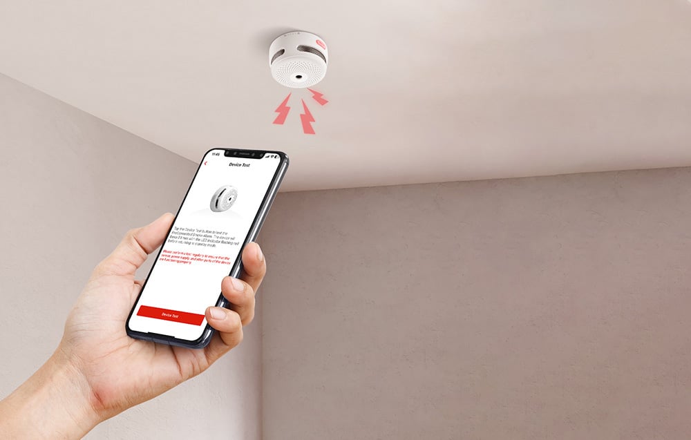 Dłoń trzymająca smartfon z aplikacją testującą czujnik dymu z czerwonymi strzałkami na suficie