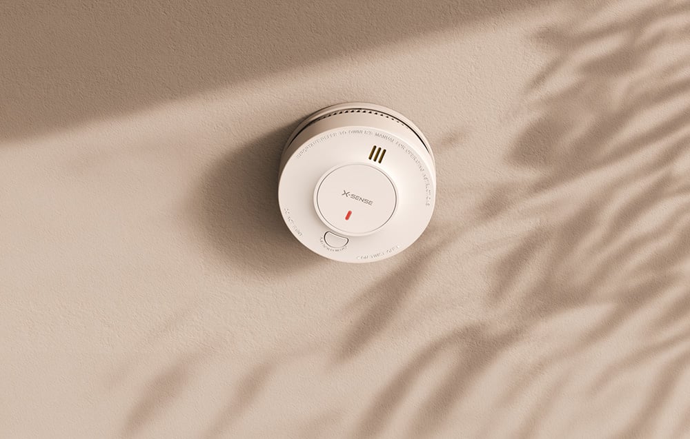 X-Sense smoke detector mounted on a beige ceiling with the shadow of a plant