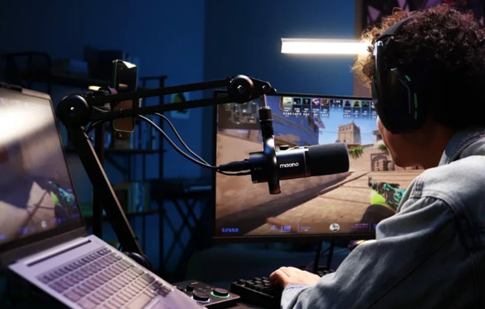 A man is playing a game, in front of him on his desk is a maono microphone