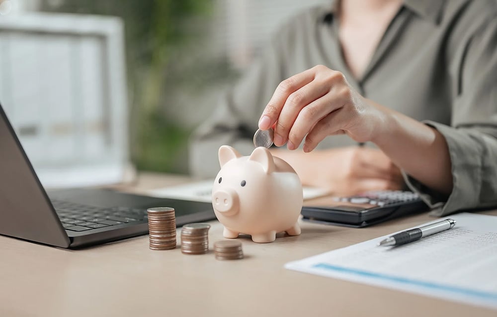 A person throwing a coin into a pink piggy bank next to a laptop and a pile of coins, symbolizing savings.