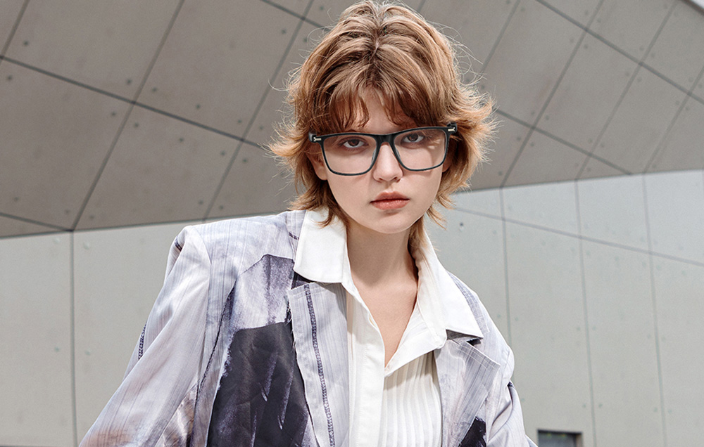 A young person in a modern blazer and black glasses poses against a concrete structure.