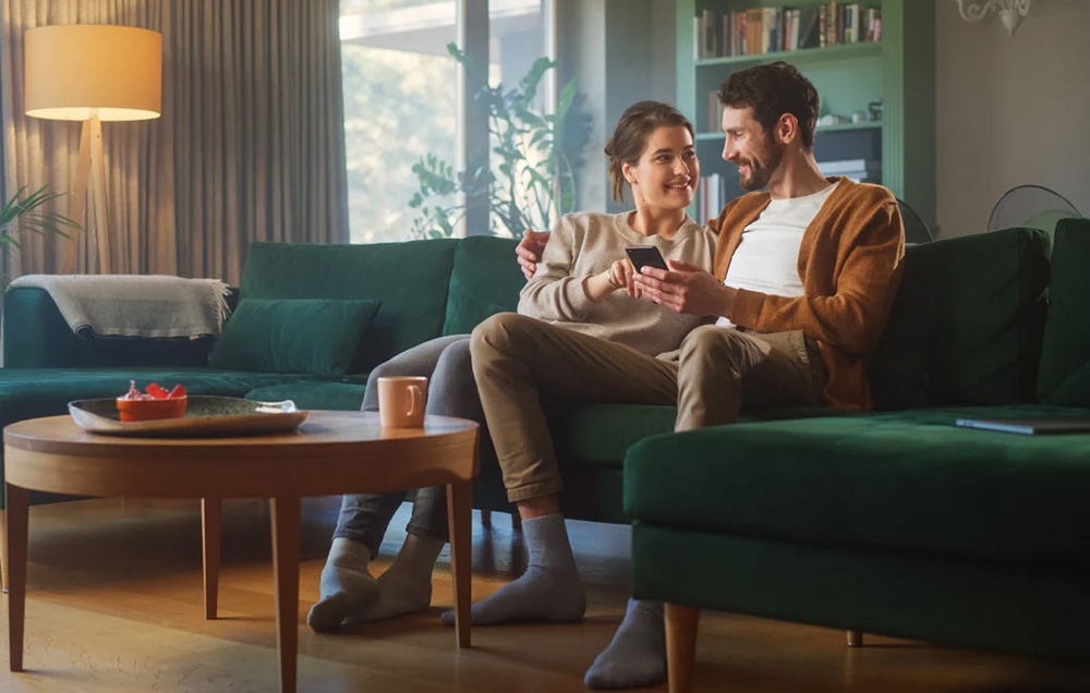 A woman and a man are sitting on a green sofa in the living room; both are looking at a phone screen, with a side table holding a cup and a bowl next to them.