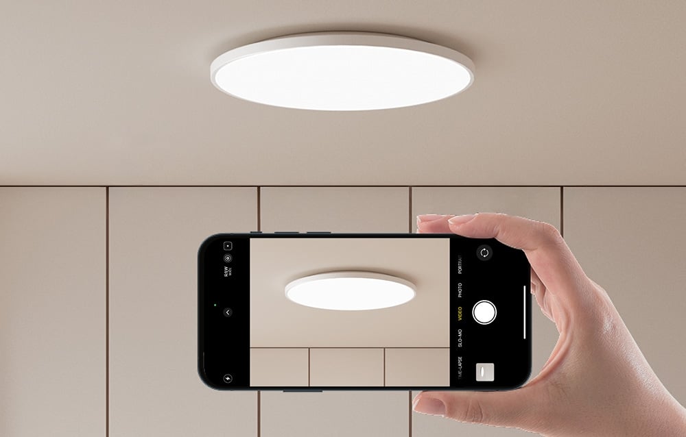 A hand holding a smartphone with the camera pointed at the Yeelight ceiling lamp on.