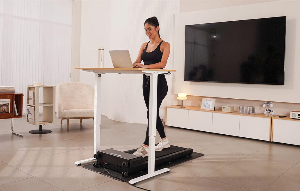 Smiling woman working on a laptop at a white desk while standing on a black treadmill