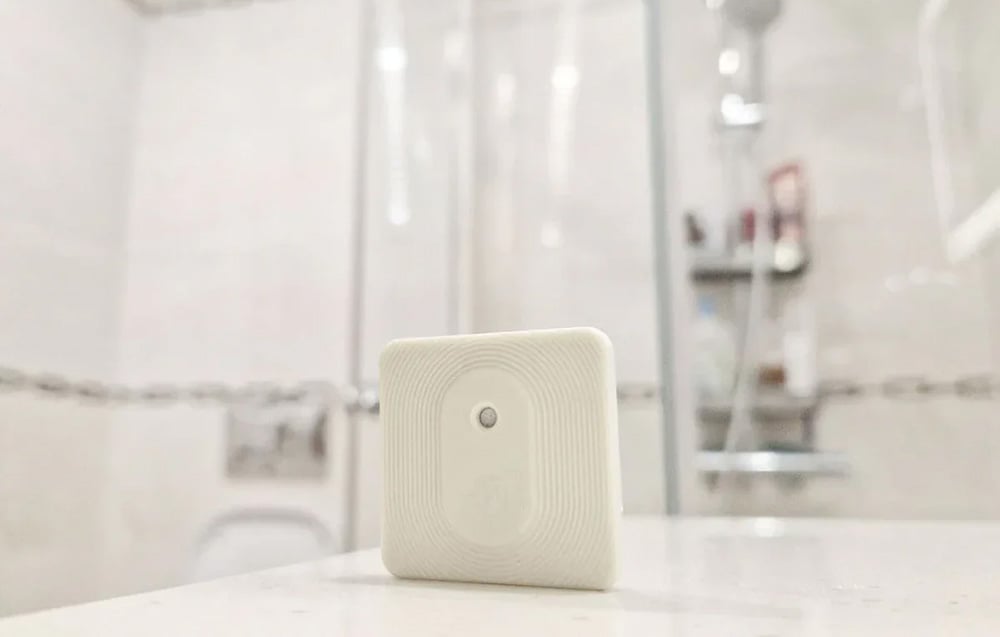 Compact Shelly temperature and humidity sensor in flesh color, placed on a countertop in a modern bathroom.