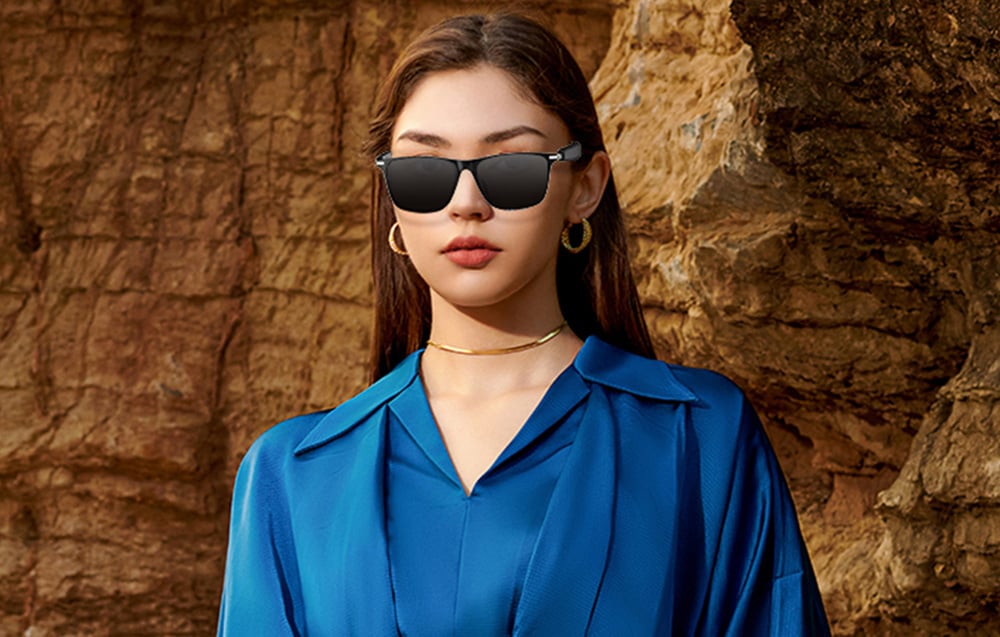 A woman in a blue blouse presenting black smart sunglasses against a backdrop of rocks.