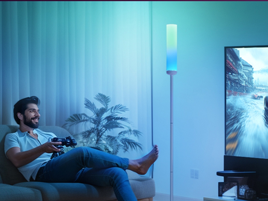 A man sitting on a couch with a game controller in front of the TV, next to a tall lamp with blue-green light.