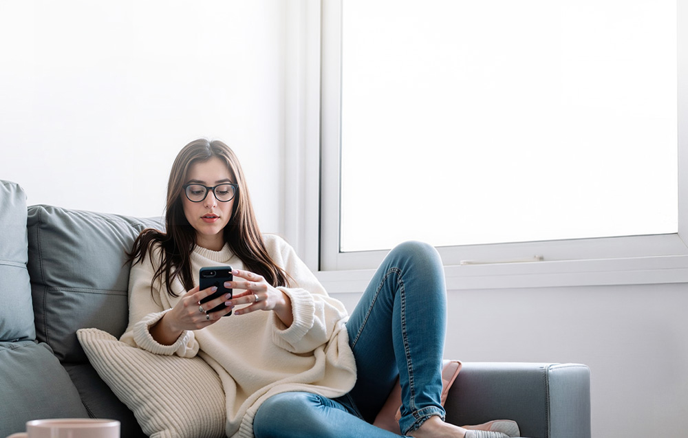 A woman wearing glasses sits on a couch holding a smartphone