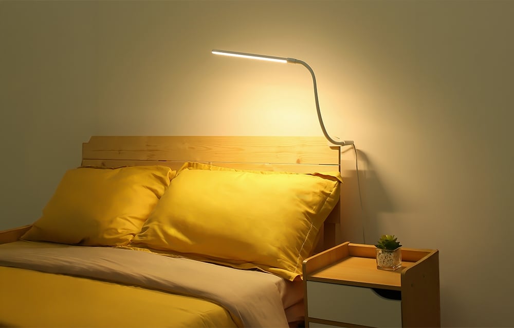 A bedroom with a wooden headboard, yellow bedding, and a nightstand, illuminated by a desk lamp mounted above.