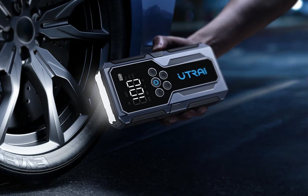 A car jump starter held in the hand illuminates the area around a car at night using its built-in flashlight.