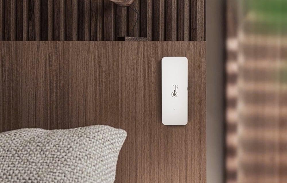Temperature and humidity sensor mounted on a wooden wall in a cozy interior with a visible cushion