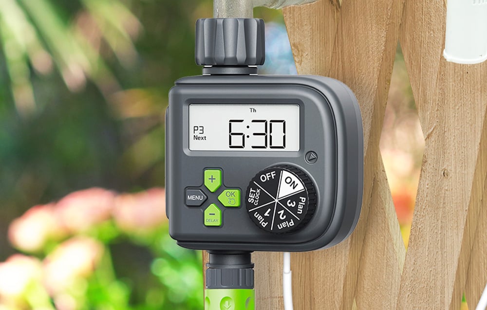 Electronic irrigation controller with a display showing 6:30, mounted on a wooden fence, with a green garden hose connected