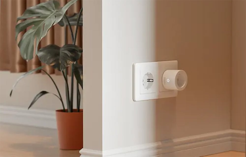 A white smart plug plugged into a wall outlet, with a potted plant and a curtain in the background