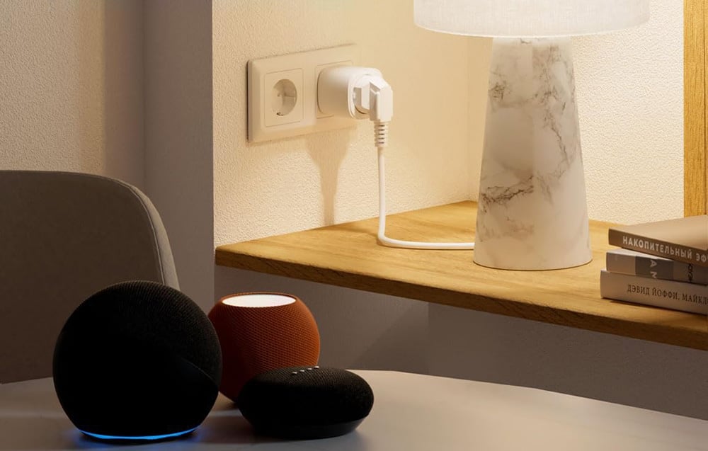 Three Amazon Echo smart speakers in black, orange, and black placed on a table in front of a smart outlet connected to a power outlet and a lamp on a wooden she