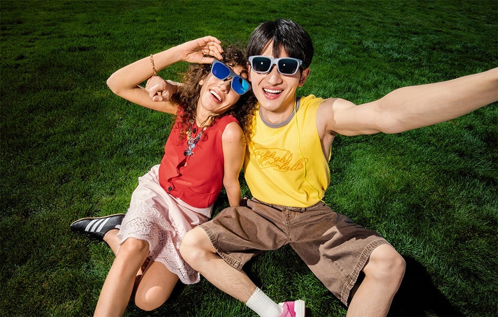 Two smiling people wearing glasses are sitting on green grass and taking a photo together from above