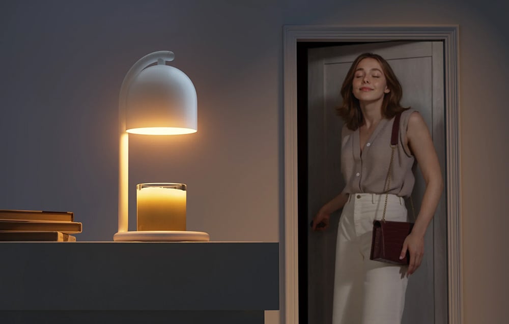 A woman with a handbag enters a darkened room through the door, where a candle warmer on a shelf is heating a large scented candle in a glass jar.