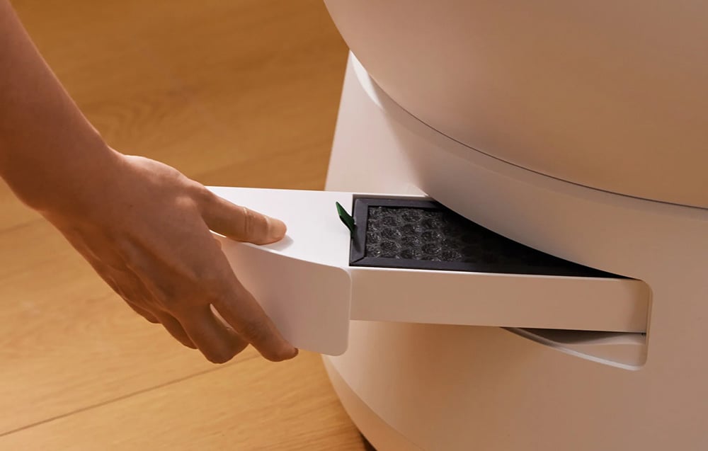 A hand pulls out a rectangular filter from the side of a white litter box standing on a wooden floor