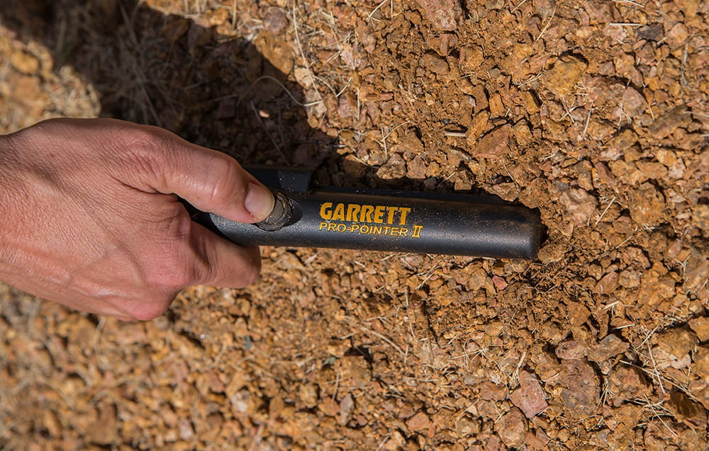A hand holds a handheld metal detector against the ground covered with dry clay.