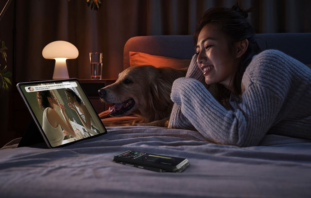 A woman lying on a bed with her dog, watching a video on a tablet; next to her, on the duvet, lies a HOTVWAV smartphone.