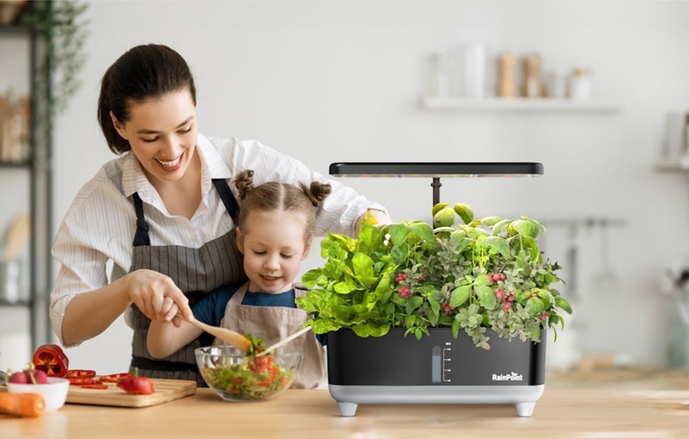 A woman and a child are preparing a salad in a bowl next to a home hydroponic garden with herbs.