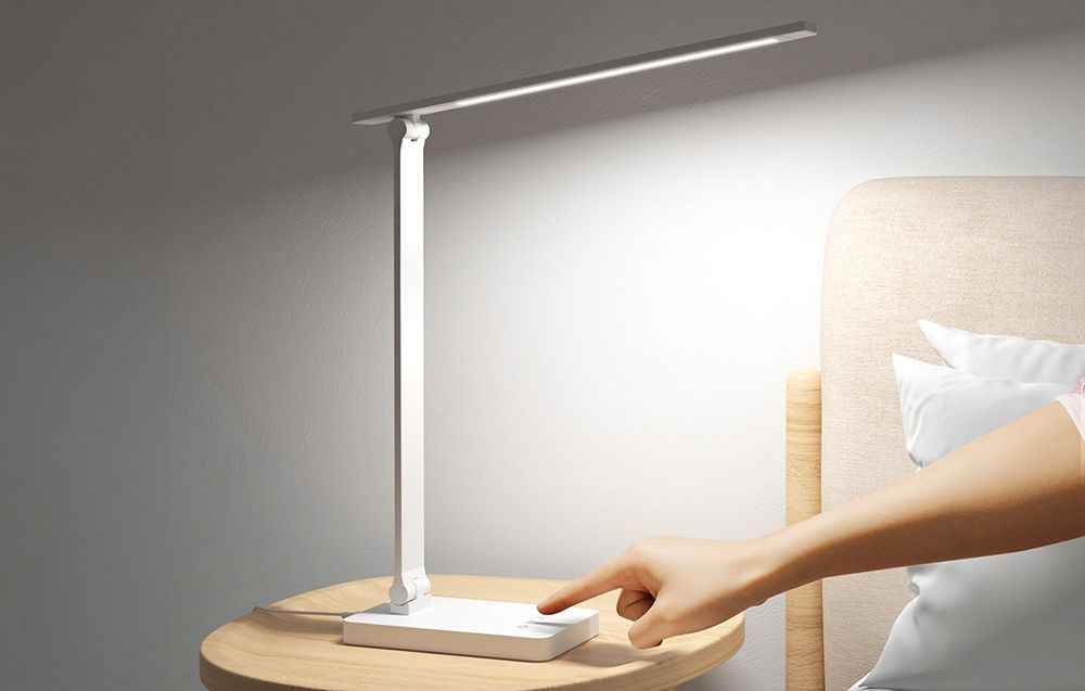 A finger touches the control panel of the lamp on the nightstand, which illuminates the space by the bed with a bright beam of light.